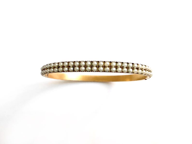 Puede incluir: Una pulsera dorada con dos filas de peque&ntilde;as perlas redondas de color blanco roto. Las perlas est&aacute;n muy juntas, creando una superficie texturizada. La pulsera est&aacute; sobre un fondo blanco.