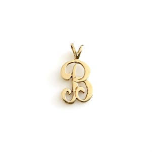 May include: A gold-colored pendant shaped like the uppercase letter B in a cursive script font. The pendant features a polished metallic finish and includes a small triangular bail at the top for attachment to a chain or necklace cord.