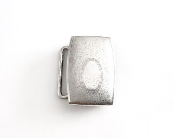 May include: A rectangular, silver-toned belt buckle with a decorative oval design in the center. The buckle has a simple, rectangular frame and a single, curved bar for the belt strap. The metal has a slightly aged appearance.