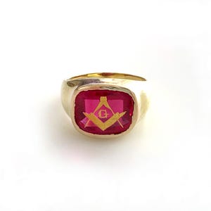 May include: A gold-toned ring with a large, rectangular, ruby-red stone. The stone features a gold-colored Masonic symbol. The ring has a classic, signet-style design, with a smooth, polished band.