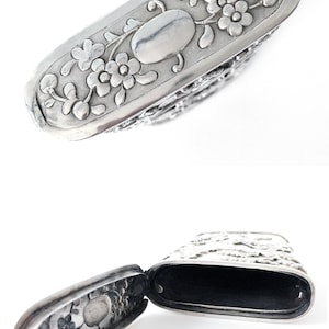 May include: A silver, hinged, rectangular box with an ornate floral design. The box is open, revealing a black interior.