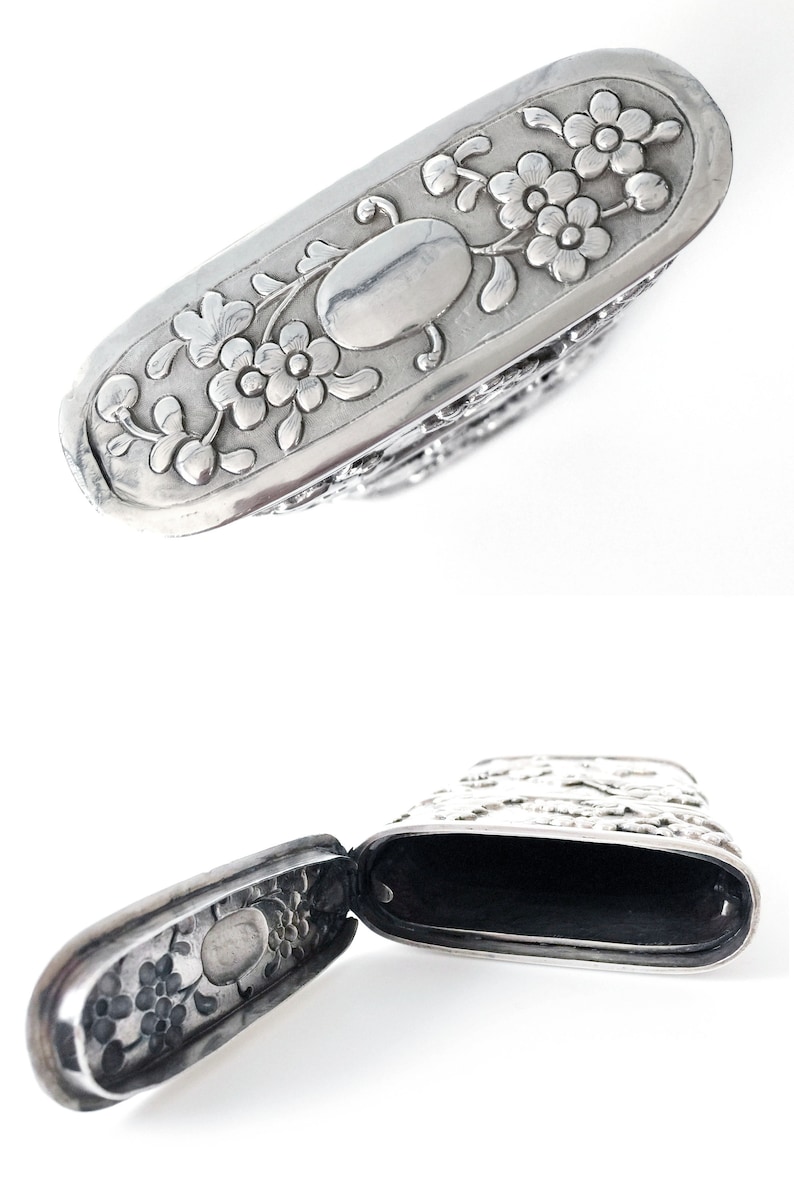 May include: A silver, hinged, rectangular box with an ornate floral design. The box is open, revealing a black interior.