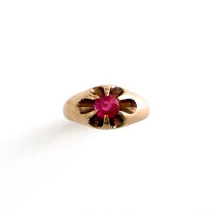 May include: A gold ring featuring a single round, vibrant pinkish-red gemstone set in the center. The metal setting around the stone is shaped like a flower with six distinct petals, creating a smooth, rounded profile for the band.