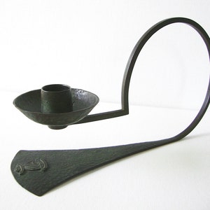 Puede incluir: Un candelero de metal verde oscuro con un brazo curvo y una base circular. La base tiene una superficie texturizada y un peque&ntilde;o dise&ntilde;o en el centro.