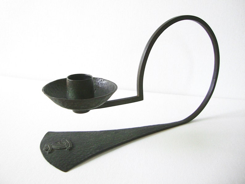 Puede incluir: Un candelero de metal verde oscuro con un brazo curvo y una base circular. La base tiene una superficie texturizada y un peque&ntilde;o dise&ntilde;o en el centro.