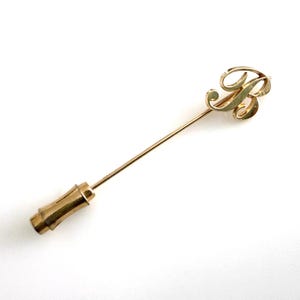 May include: A gold-toned stick pin featuring a decorative script letter R at the top. The pin has a long, slender metal shaft and a cylindrical, textured end cap that resembles a small bamboo segment, providing a secure closure for the accessory.
