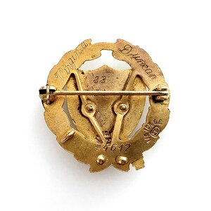 May include: A vintage gold-toned pin with a circular design. The pin features the engraved words "Virginia Duncan" and the number "83". The pin has a shield shape in the center and a pin clasp on the back. The pin is on a white background.