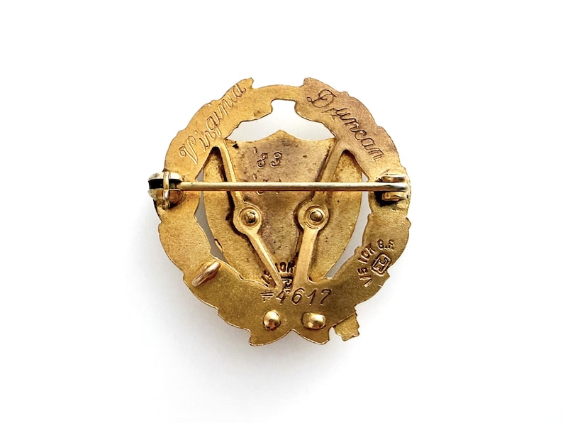 May include: A vintage gold-toned pin with a circular design. The pin features the engraved words "Virginia Duncan" and the number "83". The pin has a shield shape in the center and a pin clasp on the back. The pin is on a white background.