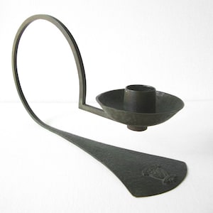 Puede incluir: Un candelero de bronce con un brazo curvo y una base redonda martillada. El candelero tiene un dise&ntilde;o simple y elegante.