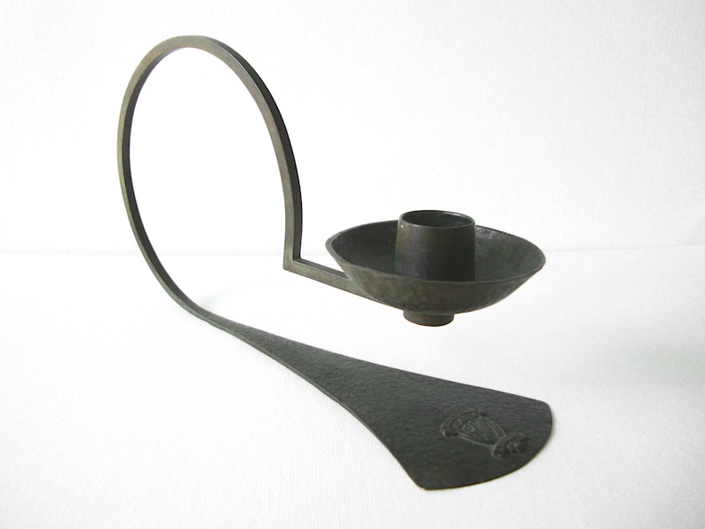 Puede incluir: Un candelero de bronce con un brazo curvo y una base redonda martillada. El candelero tiene un dise&ntilde;o simple y elegante.