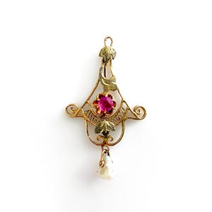 May include: A gold pendant featuring a central round pink gemstone set within a filigree frame. The design includes engraved leaf motifs at the top and bottom, with a small, irregular white pearl drop hanging from the base of the structure.