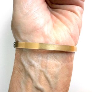 Puede incluir: Un brazalete rectangular de color dorado con un acabado cepillado. El brazalete se lleva en la mu&ntilde;eca y tiene un dise&ntilde;o simple y elegante. El brazalete mide aproximadamente 1,3 cm de ancho.