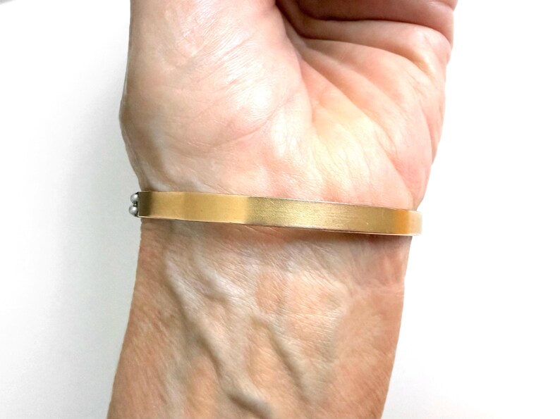 Puede incluir: Un brazalete rectangular de color dorado con un acabado cepillado. El brazalete se lleva en la mu&ntilde;eca y tiene un dise&ntilde;o simple y elegante. El brazalete mide aproximadamente 1,3 cm de ancho.