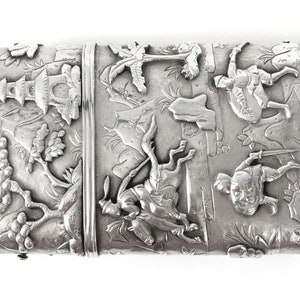 May include: A silver rectangular box with an intricate design of Asian figures and scenes. The box is decorated with raised details and has a hinged lid.