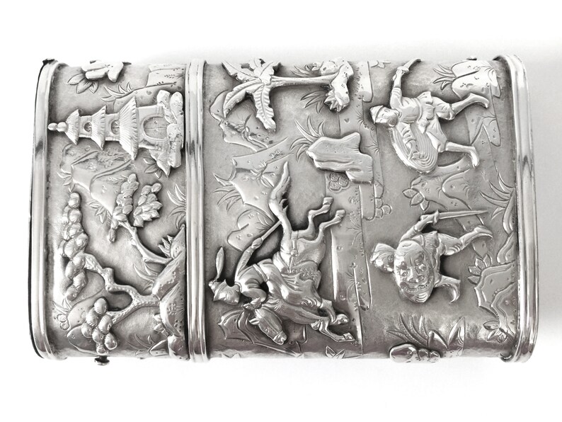 May include: A silver rectangular box with an intricate design of Asian figures and scenes. The box is decorated with raised details and has a hinged lid.