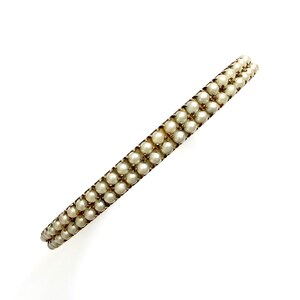 Puede incluir: Una pulsera de estilo vintage con dos filas de peque&ntilde;as perlas redondas de color blanco roto. Las perlas est&aacute;n engarzadas en un marco de metal dorado. La pulsera se presenta sobre un fondo blanco liso, resaltando su elegante dise&ntilde;o.