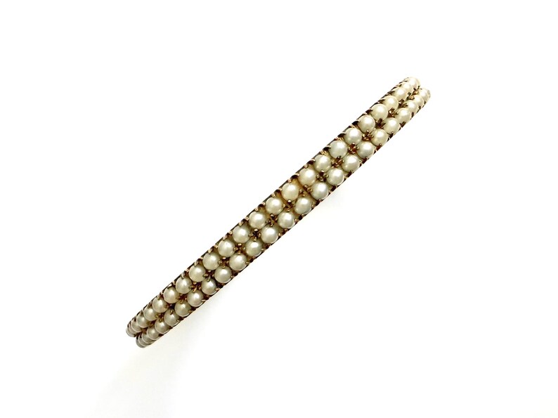 Puede incluir: Una pulsera de estilo vintage con dos filas de peque&ntilde;as perlas redondas de color blanco roto. Las perlas est&aacute;n engarzadas en un marco de metal dorado. La pulsera se presenta sobre un fondo blanco liso, resaltando su elegante dise&ntilde;o.