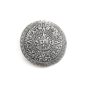 May include: A silver-colored, circular pendant featuring an intricate Aztec calendar design. The pendant has detailed carvings of symbols and figures, with a central face and surrounding patterns. The background is plain white.