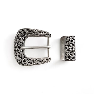 May include: A silver-toned metal belt buckle and matching rectangular keeper loop. Both pieces feature an intricate, raised filigree scrollwork pattern with a dark, oxidized finish in the recessed areas, providing a high-contrast, textured appearance.