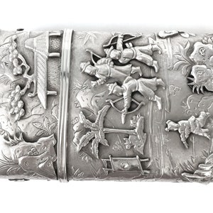 May include: A silver rectangular box with an intricate design of people with bows and arrows, trees, and a building. The box is decorated with a raised design.