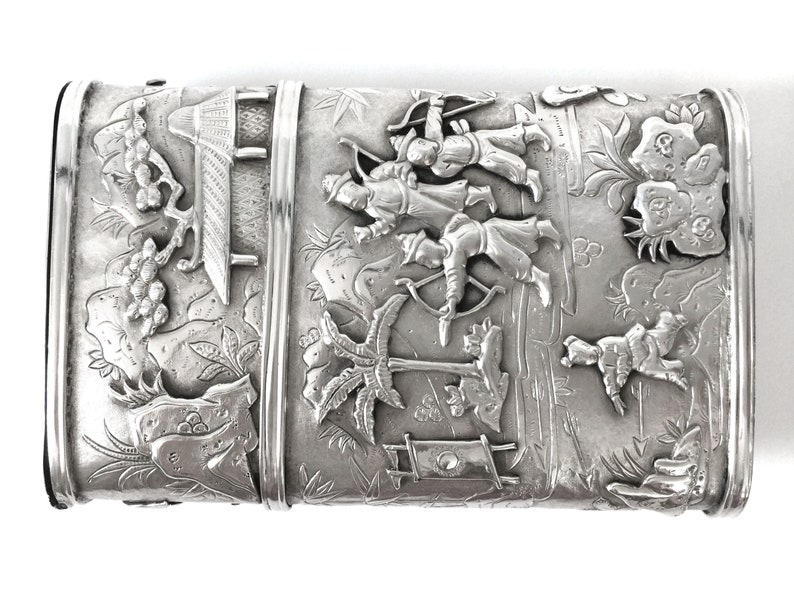 May include: A silver rectangular box with an intricate design of people with bows and arrows, trees, and a building. The box is decorated with a raised design.