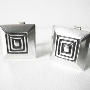 Può includere: Un paio di gemelli d'argento con un design quadrato. Ogni gemello presenta un motivo quadrato concentrico con un centro rialzato.