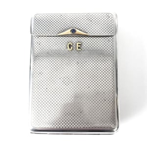 May include: A silver cigarette case with a checkered pattern and a gold-colored hinge. The initials "CE" are engraved on the front.