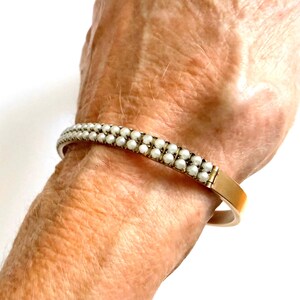 Puede incluir: Un brazalete dorado con dos filas de peque&ntilde;as perlas blancas. El brazalete se muestra en una mu&ntilde;eca, resaltando el dise&ntilde;o y el contraste de color. Las perlas est&aacute;n espaciadas uniformemente, creando un aspecto cl&aacute;sico y elegante.