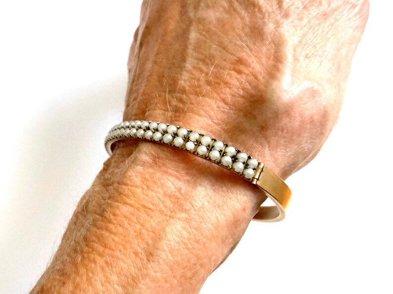 Puede incluir: Un brazalete dorado con dos filas de peque&ntilde;as perlas blancas. El brazalete se muestra en una mu&ntilde;eca, resaltando el dise&ntilde;o y el contraste de color. Las perlas est&aacute;n espaciadas uniformemente, creando un aspecto cl&aacute;sico y elegante.