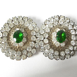 May include: A pair of vintage brooches with a circular design. Each brooch features a central green stone surrounded by a ring of smaller clear rhinestones.