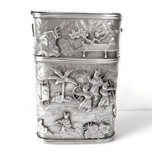 May include: A silver rectangular box with intricate embossed designs of figures and trees. The box is divided into two sections.