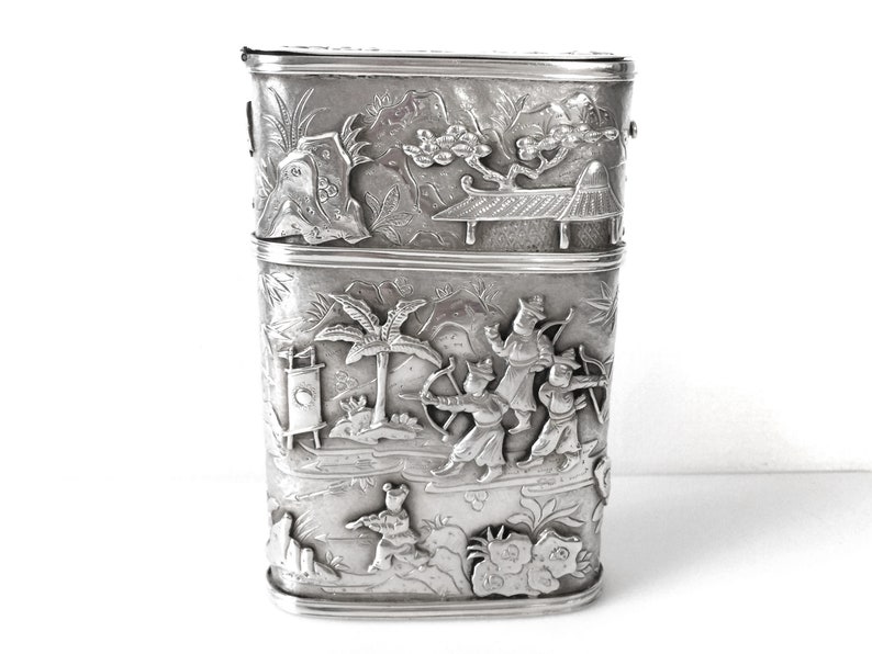 May include: A silver rectangular box with intricate embossed designs of figures and trees. The box is divided into two sections.