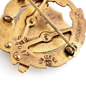 May include: Close-up of a vintage gold-toned brooch. The brooch features a central design with a shield and a laurel wreath. The pin has the text "Duncan" and "1/5 10K G.F." stamped on it. The brooch is likely made of gold-filled metal.