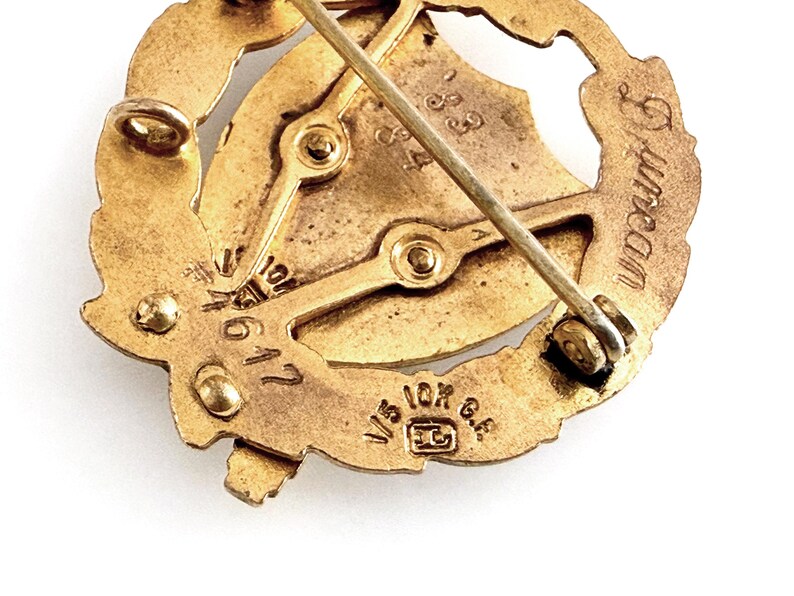 May include: Close-up of a vintage gold-toned brooch. The brooch features a central design with a shield and a laurel wreath. The pin has the text "Duncan" and "1/5 10K G.F." stamped on it. The brooch is likely made of gold-filled metal.