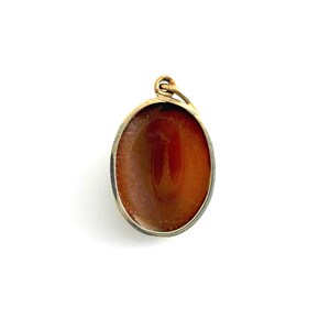 K&ouml;nnte beinhalten: Ovaler Anh&auml;nger mit einem polierten, rotbraunen Stein, der in einen goldfarbenen Metallrahmen gefasst ist. Der Anh&auml;nger hat oben eine Schlaufe f&uuml;r eine Kette. Der Stein hat eine gl&auml;nzende Oberfl&auml;che.