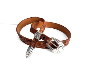 May include: A brown leather belt featuring an intricate floral embossed pattern throughout the strap. The belt is equipped with a polished silver-tone metal buckle, a matching silver-tone belt tip, and a silver-tone metal keeper loop.