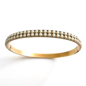 Puede incluir: Un brazalete dorado con dos filas de peque&ntilde;as perlas redondas de color blanquecino. El brazalete tiene un cierre de bisagra y un acabado liso y pulido, sobre un fondo blanco.