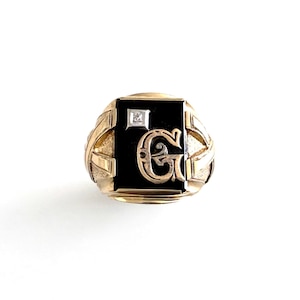 Op de afbeelding: Een gouden ring met een zwart onyx middenstuk met een diamant en de letter "G" in een decoratief schrift. De ring heeft een rechthoekige voorkant en gedetailleerd goudwerk aan de zijkanten.