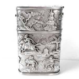 May include: A silver rectangular box with intricate embossed designs of Asian figures and landscapes. The box is divided into two sections.