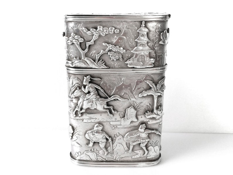 May include: A silver rectangular box with intricate embossed designs of Asian figures and landscapes. The box is divided into two sections.