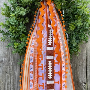 May include: A wreath with orange and white ribbons, a football, and a pom-pom trim. The ribbons have the words "Tennessee" and "Volunteers" printed on them.