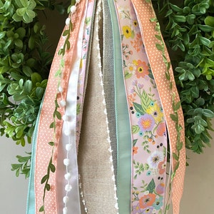 May include: A decorative wreath with a lush green base, adorned with various ribbons. The ribbons feature floral patterns, polka dots, and solid colors like peach and mint green. Some ribbons have white pom-poms and delicate leaf accents.