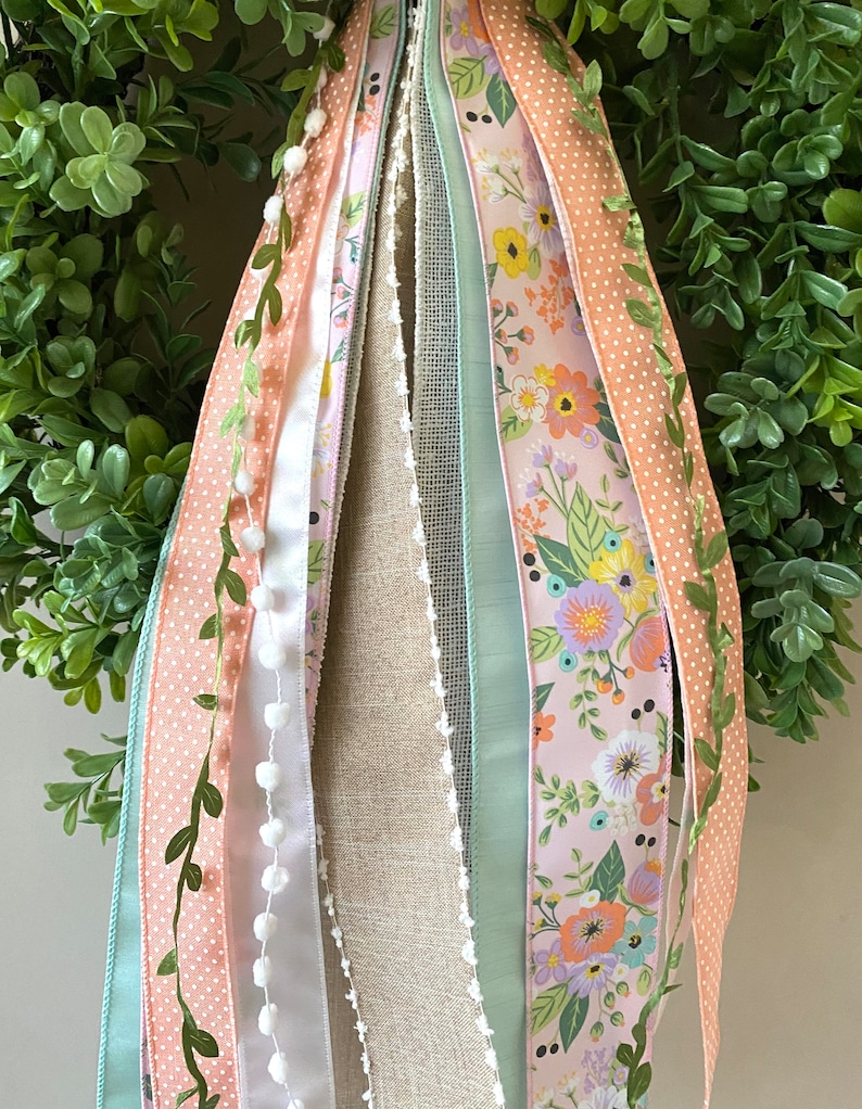 May include: A decorative wreath with a lush green base, adorned with various ribbons. The ribbons feature floral patterns, polka dots, and solid colors like peach and mint green. Some ribbons have white pom-poms and delicate leaf accents.
