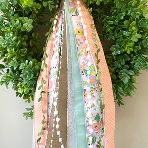 May include: A green, leafy wreath with a variety of decorative ribbons. The ribbons include floral patterns, polka dots, and solid colors in shades of pink, peach, and green. A string of white pom-poms adds a delicate touch.