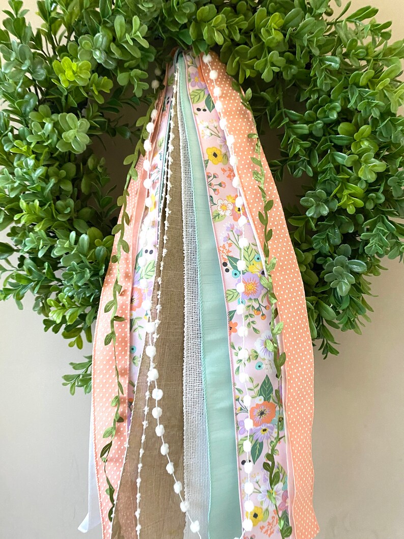 May include: A green, leafy wreath with a variety of decorative ribbons. The ribbons include floral patterns, polka dots, and solid colors in shades of pink, peach, and green. A string of white pom-poms adds a delicate touch.