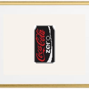 以下が含まれることがあります： 額縁に入ったクロスステッチアート。コカ・コーラ ゼロの缶が描かれています。缶は黒色で、コカ・コーラのロゴは赤色、"zero"の文字は白色です。金色のフレームと白いマットで飾られています。