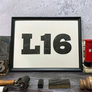 May include: Framed print featuring the letters "L16" in black, set against a white backdrop. The image is surrounded by printing tools, including a roller, stamps, and a miniature red postbox.