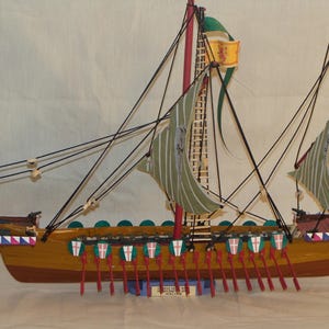 May include: A wooden model of a medieval galley ship with red and white striped sails, green and white striped sails, and a red and white striped flag. The ship has a wooden hull with red and white stripes and a row of red oars.