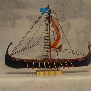 May include: A model of a Viking longship with a dragon head prow, a blue and orange sail, and a wooden mast. The ship is decorated with shields and oars.
