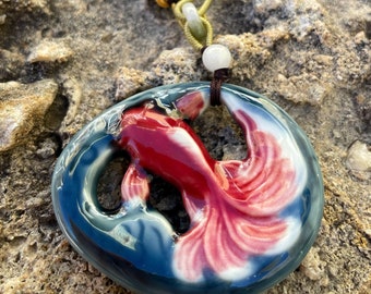 May include: A necklace with a ceramic pendant shaped like a red and white goldfish. The necklace is made of brown and green cord with white and orange beads.